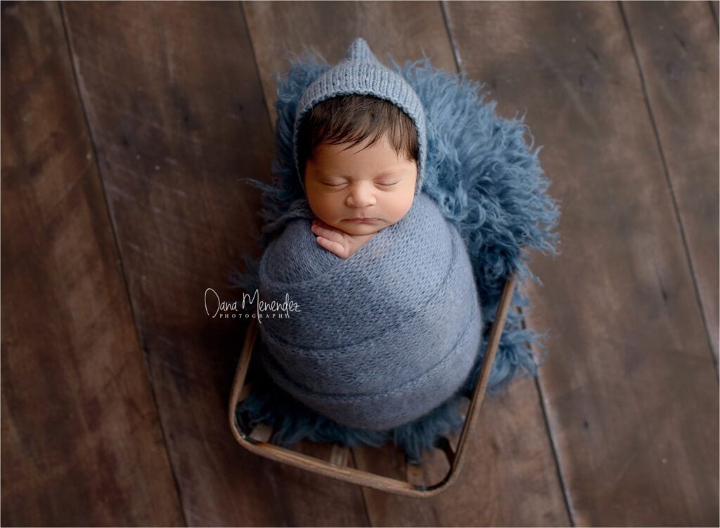 Adorable newborn wrapped in blue with matching hat, peacefully sleeping on a wooden floor.