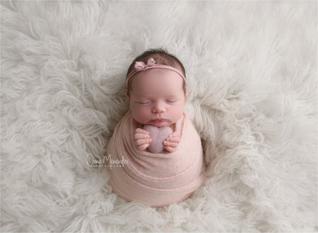 Baby girl wrapped in pink blanket with headband, sleeping peacefully on fluffy white background.