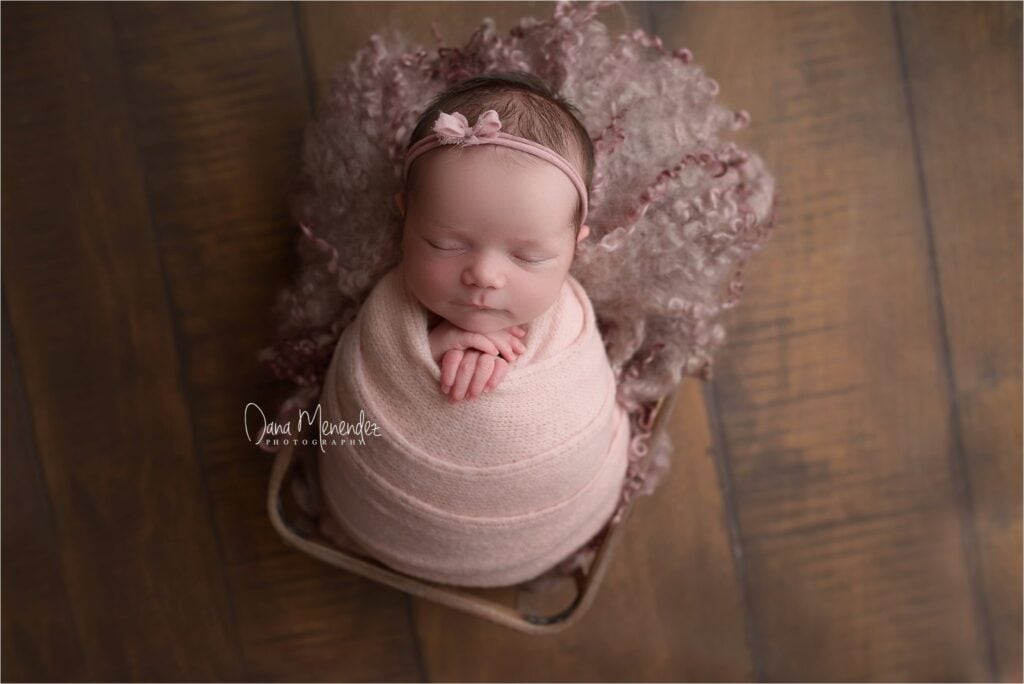 Adorable newborn girl wrapped in pink, sleeping peacefully on a wooden floor.