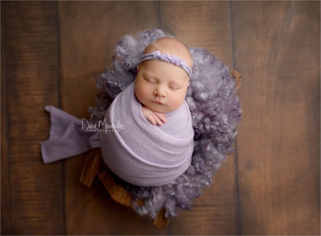 Baby wrapped in lavender swaddle with headband, sleeping peacefully.