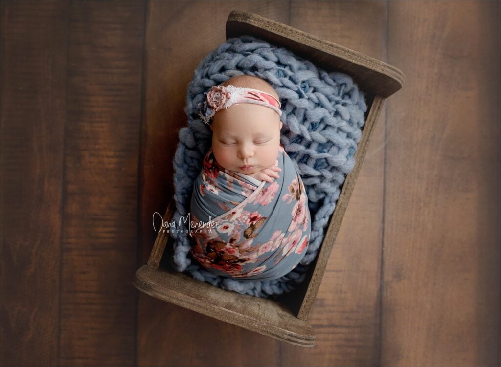 Baby girl wrapped in floral blanket with headband, sleeping peacefully in a rustic wooden box.