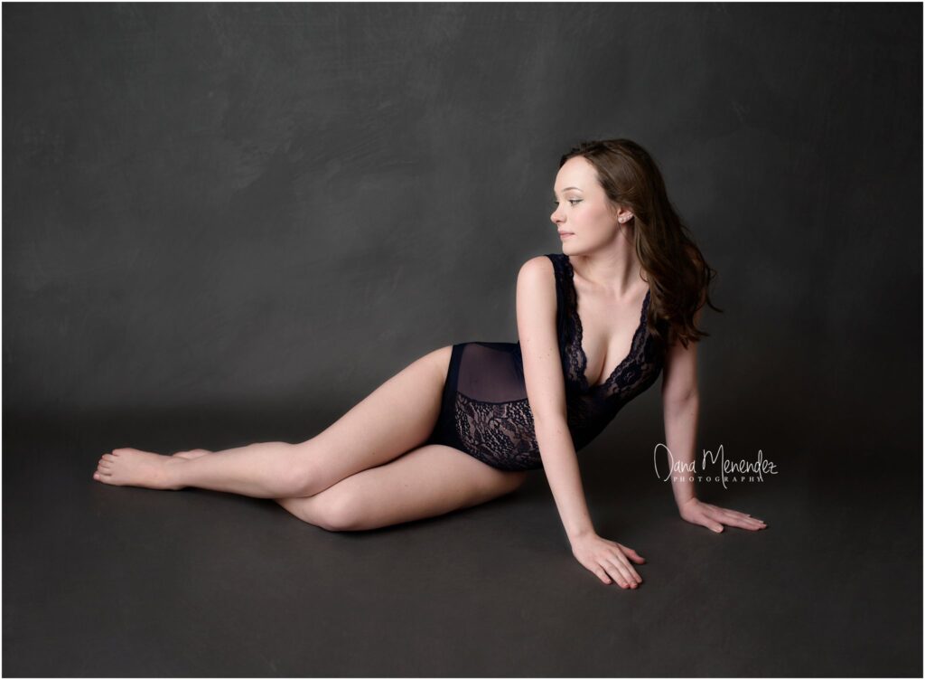A woman in black lace lingerie lying on a dark gray background, posing elegantly for a professional.
