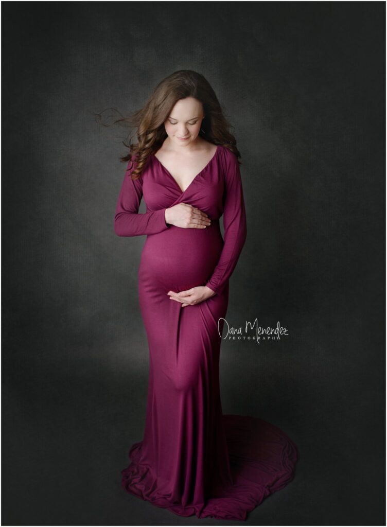Pregnant woman in a deep magenta gown holding her belly, smiling softly against a dark background.