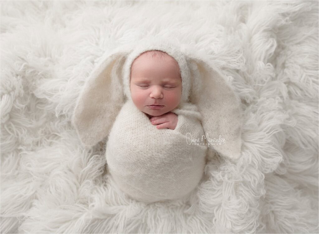 Adorable newborn baby wrapped in cream swaddle on fluffy white blanket.