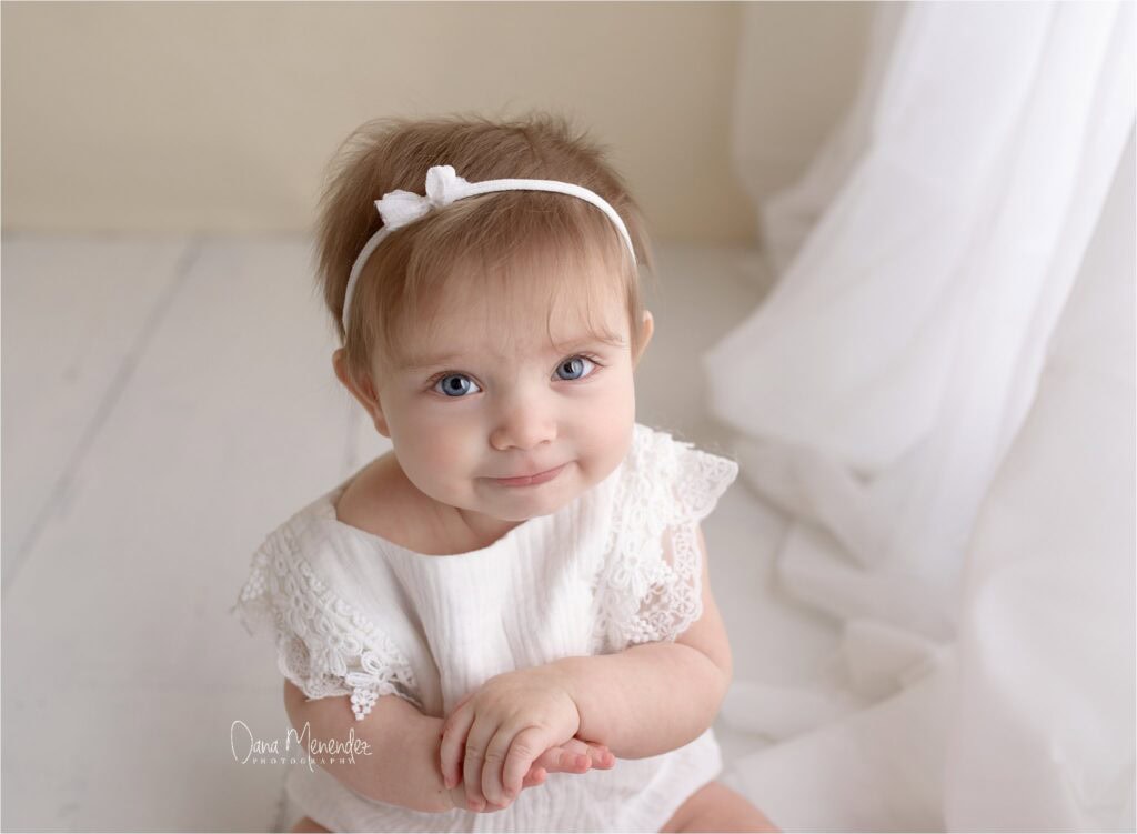 Cute newborn girl with blue eyes and a white headband, sitting on a light-colored floor.