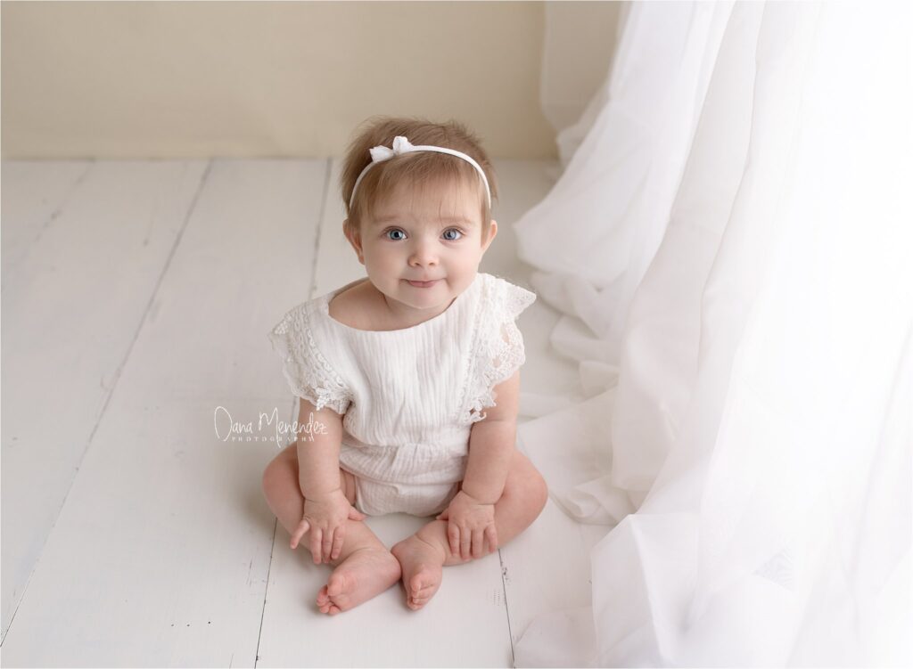 Baby girl sitting on the floor with a white dress and headband.