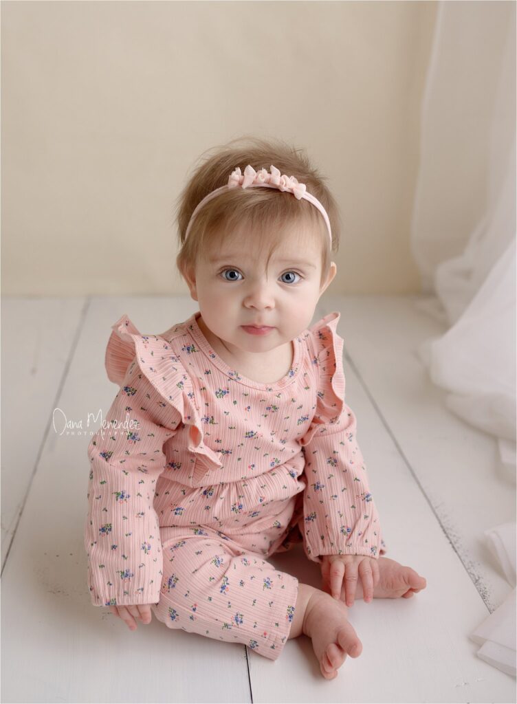 Newborn girl in pink floral outfit with headband sitting on white floor.