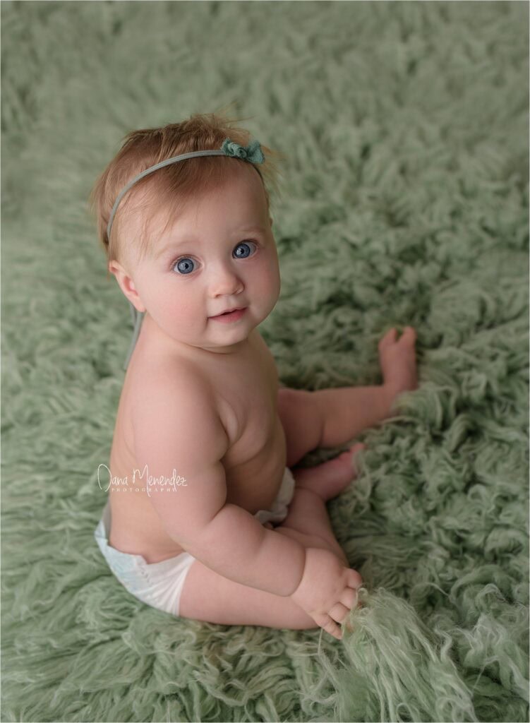 Adorable baby girl with blue eyes sitting on a soft green fuzzy blanket.