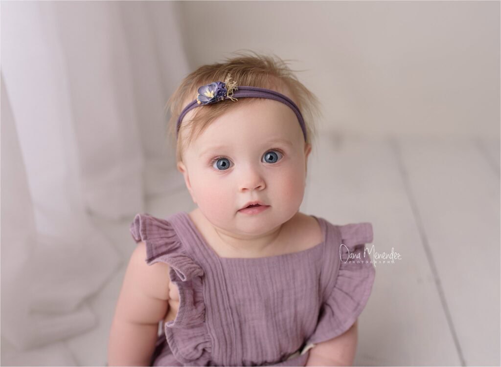 Adorable baby girl with blue eyes wearing a purple dress and headband.