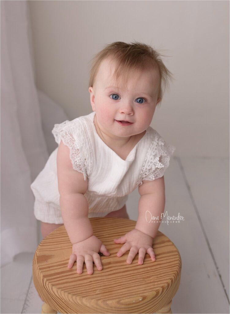 Baby girl with blue eyes and light hair, wearing a white lace dress, leaning forward on a wooden sto.