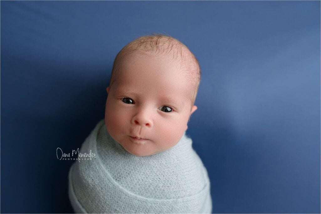 Baby portrait of a newborn wrapped in a light blue blanket against a blue background.
