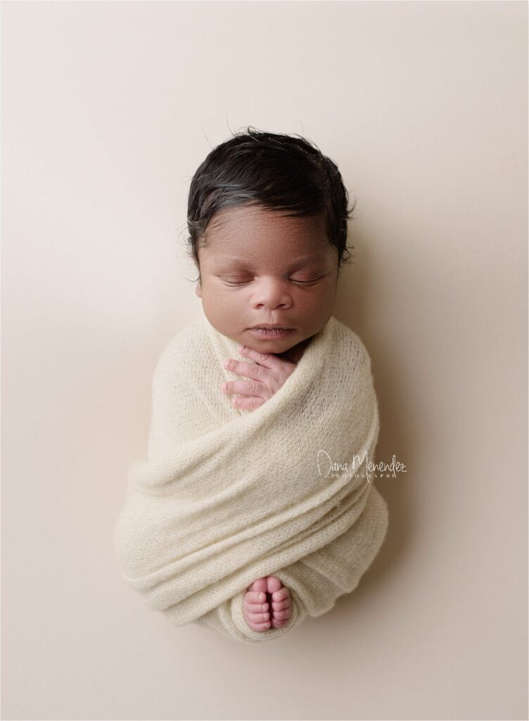 Newborn baby peacefully sleeping wrapped in a cream blanket against a neutral background.