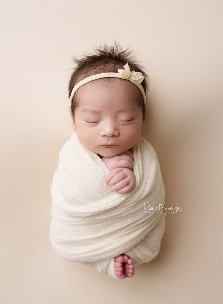 Adorable newborn girl wrapped in a soft cream blanket with a headband, peacefully sleeping.