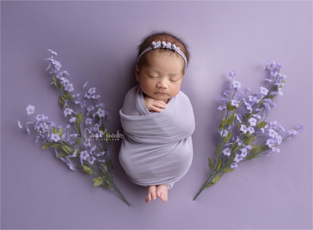 Adorable newborn girl wrapped in gray, lying on purple background with lavender flowers.