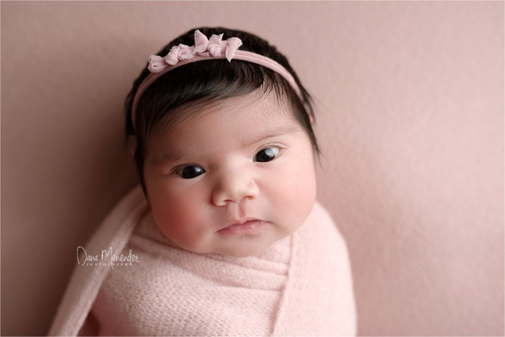 Baby girl wrapped in a pink blanket with a matching headband, looking softly at the camera.