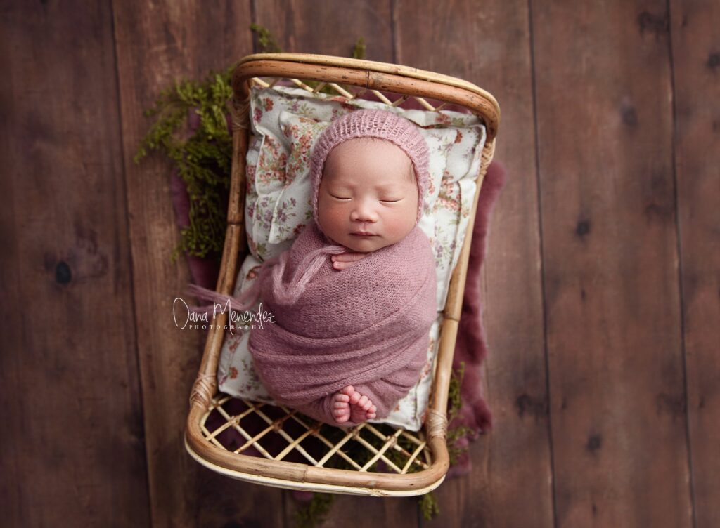 Adorable newborn baby sleeping in a wicker basket with pink wrap and hat.