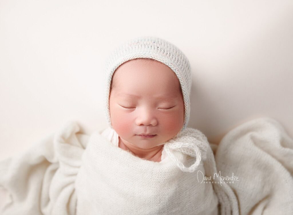 Newborn baby peacefully sleeping in a white knit hat and cozy blanket.