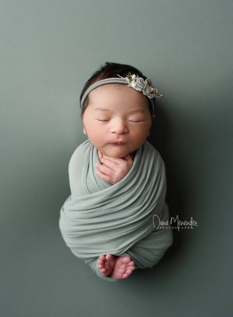 Adorable newborn girl wrapped in a soft green swaddle with a floral headband.