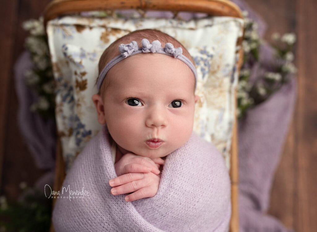 Adorable newborn girl wrapped in lavender blanket with floral background.
