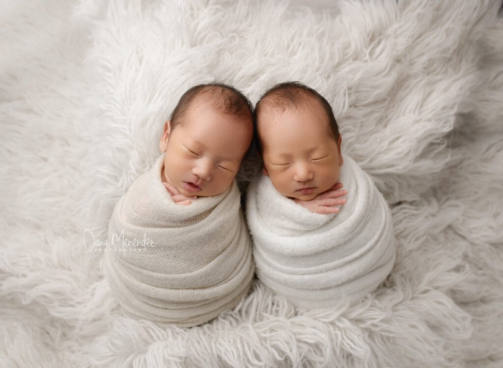 Newborn twins wrapped in soft blankets, peacefully sleeping on fluffy white fur background.