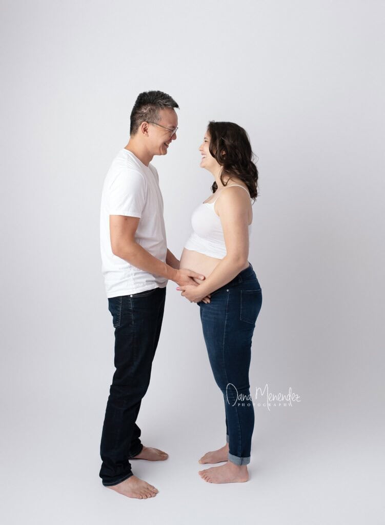 Pregnant woman and partner holding hands, smiling, and looking at each other against a plain white b.