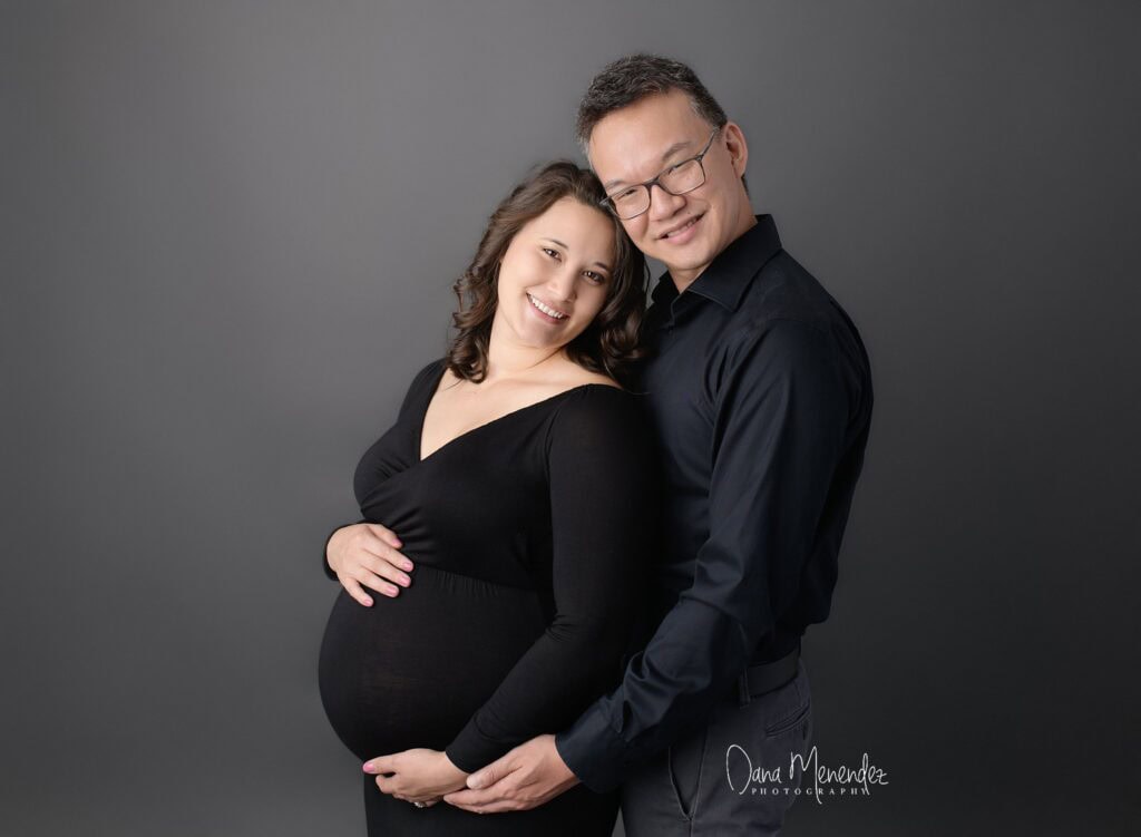 Pregnant woman and partner smiling, embracing against gray background.