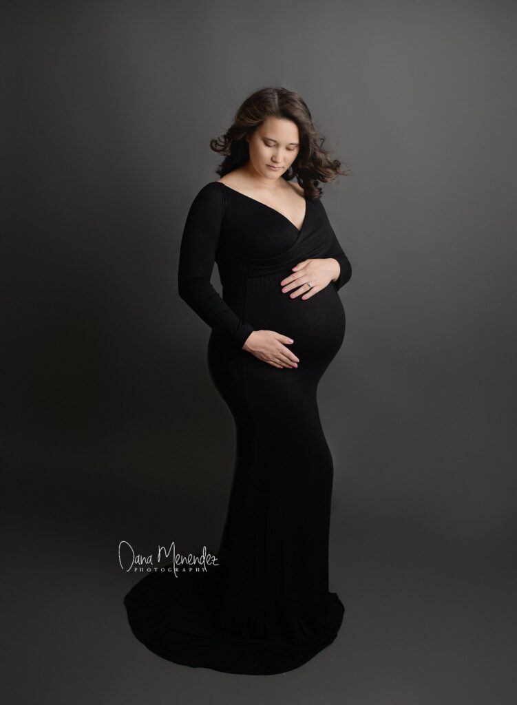 Beautiful pregnant woman in a black dress holding her belly against a gray background.