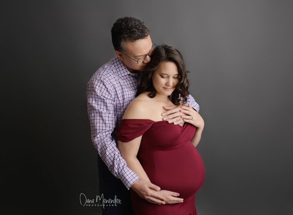 Pregnant woman with partner embracing her belly against a dark background.