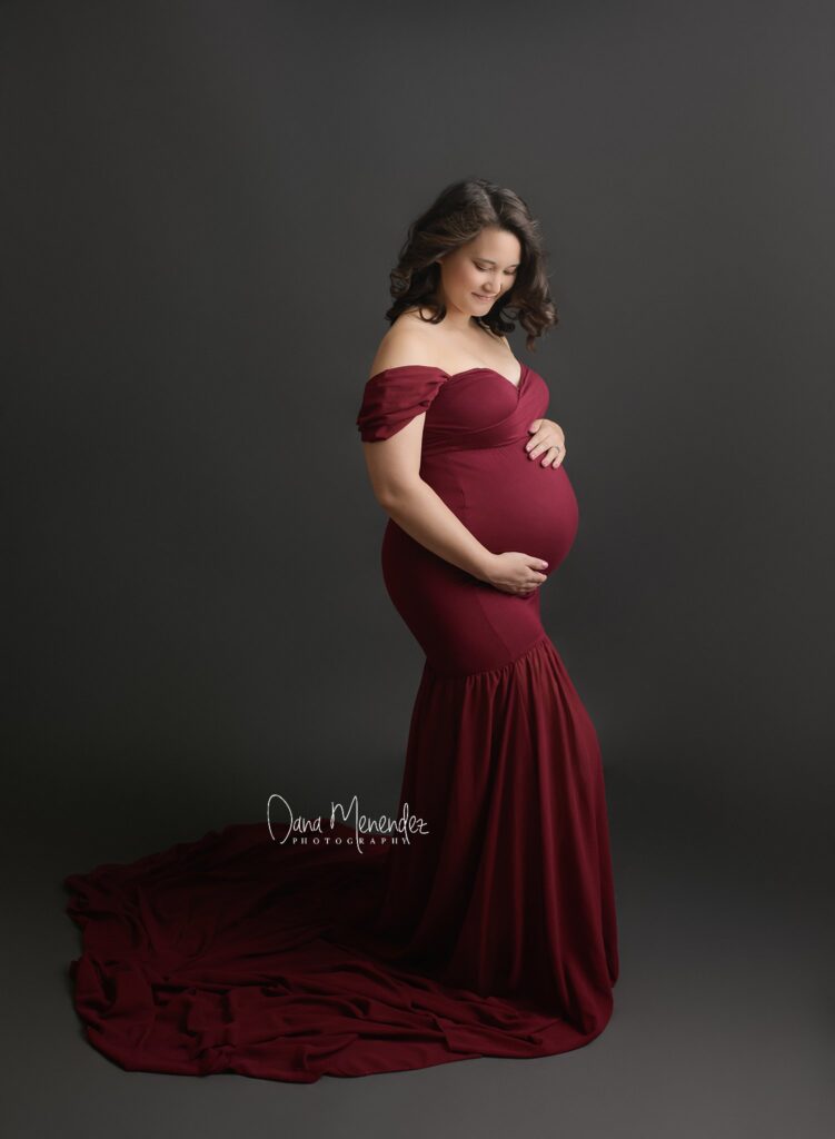 Pregnant woman in a burgundy off-shoulder gown, gently holding her belly, standing against a dark gr.