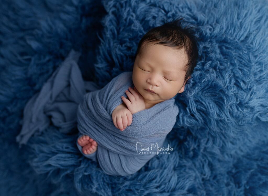 Newborn baby sleeping peacefully wrapped in a blue blanket on a soft, textured blue background.