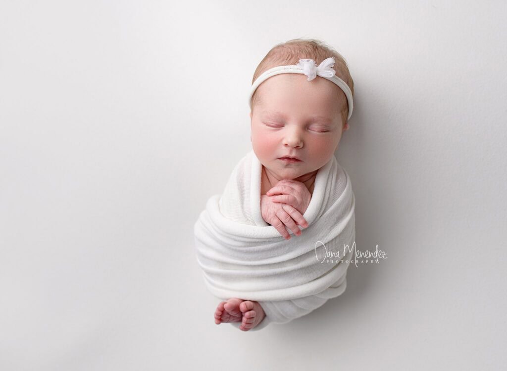 Newborn baby girl peacefully sleeping, wrapped in a white blanket with a matching headband.