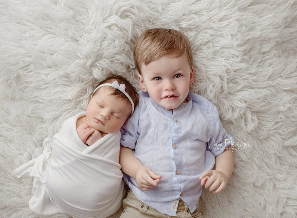 Cute newborn and toddler siblings lying on a fluffy white blanket.