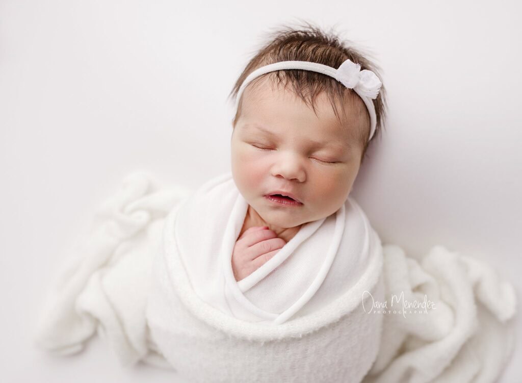 Newborn baby peacefully sleeping in a white wrap with a headband.