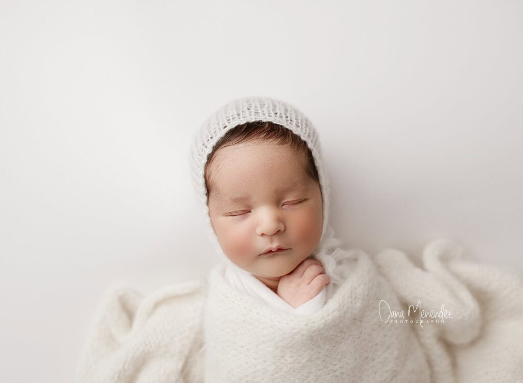 Newborn baby peacefully sleeping in a white wrap and knit hat, captured by Dana Menendez.