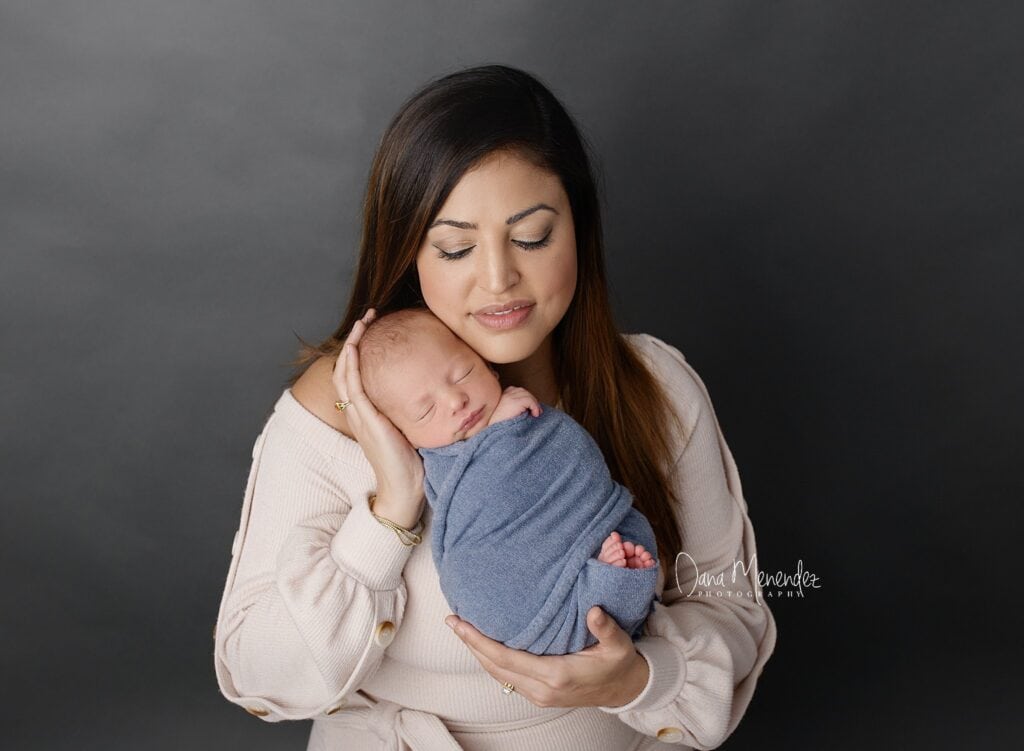 Newborn baby wrapped in blue held by mother against gray background.