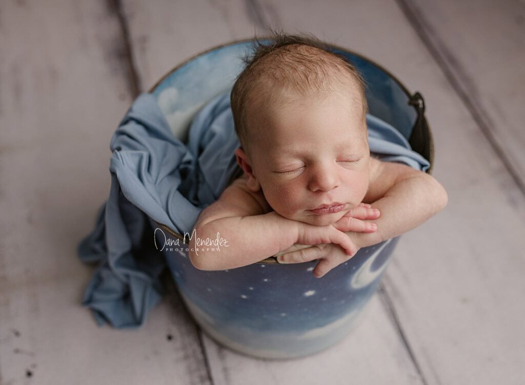 Baby sleeping in a blue bucket with a soft blanket, showcasing a peaceful newborn portrait.