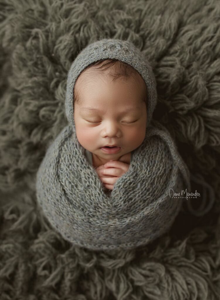 Adorable newborn baby peacefully sleeping in a gray wrap and hat on a soft textured blanket.