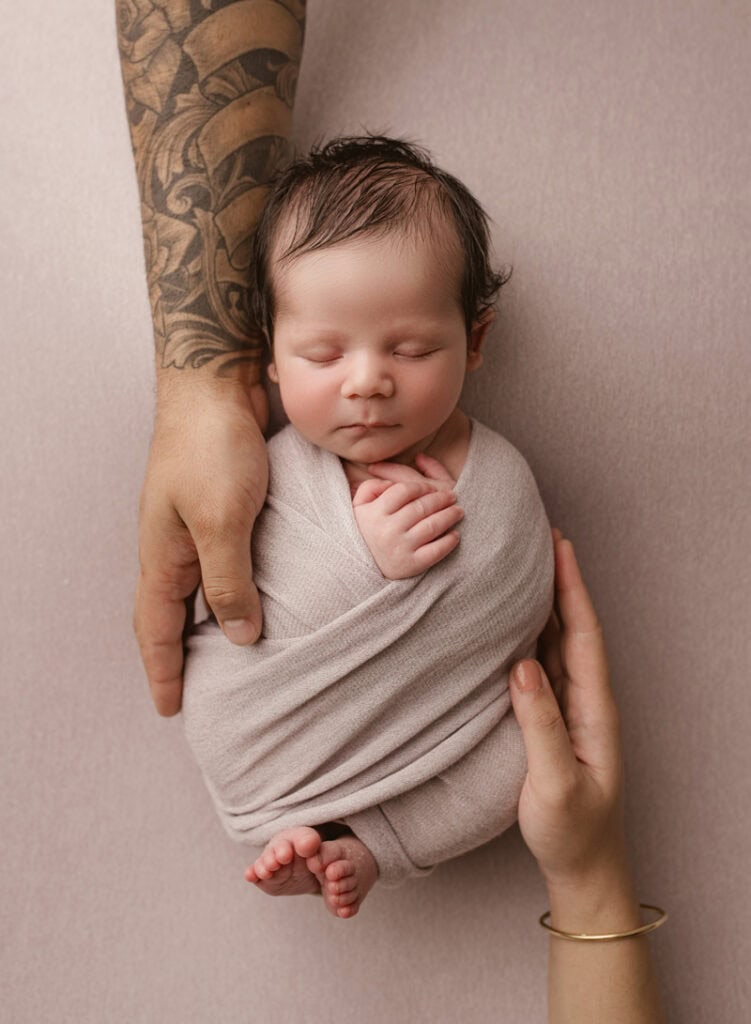 Adorable newborn baby wrapped in a blanket with hands and arm of a tattooed adult and a woman’s hand.