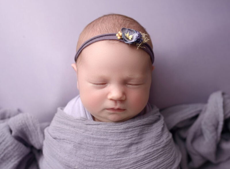Adorable newborn girl peacefully sleeping in gray wrap with floral headband.
