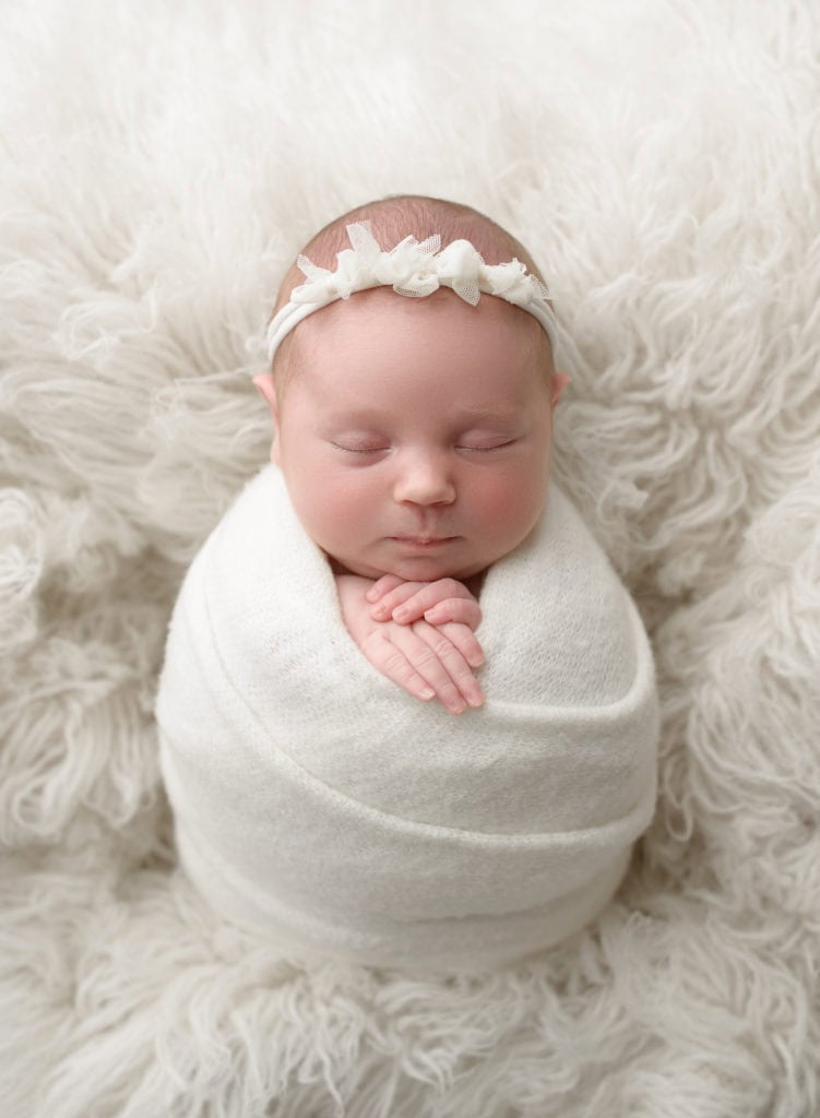 Newborn baby girl wrapped in white blanket with floral headband.