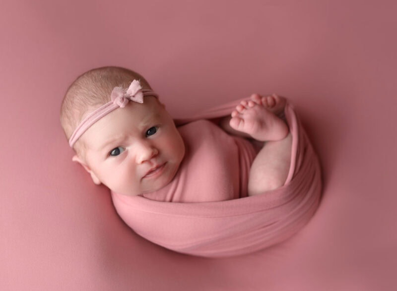Newborn baby wrapped in pink blanket with headband, cozy portrait.